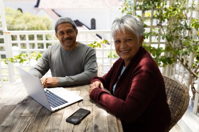 Senior African American couple sitting on a terrace, using a laptop and smiling at the camera. Ideal for content related to retirement lifestyle, technology use among seniors, self-isolation during the COVID-19 pandemic, and promoting digital literacy in older adults.