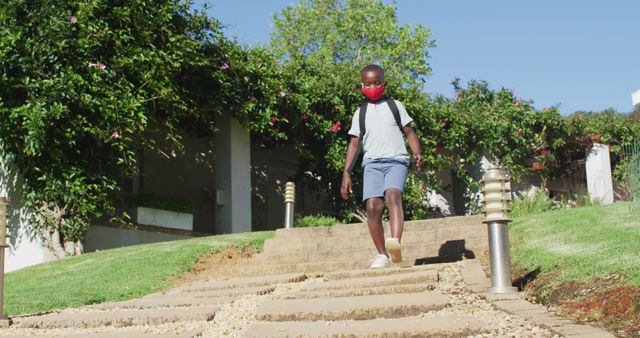 Young boy walking down garden steps while wearing a red face mask and backpack. Ideal for concepts of child safety, health precautions, COVID-19, summer activities, and outdoor exploration in nature.