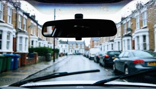 Rearview mirror framing rain-soaked windshield over parked cars on urban terraced street