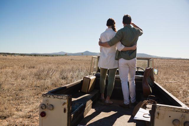 Young couple standing on the back of an off-road vehicle, embracing while looking at the open field and distant mountains. Ideal for themes related to travel, adventure, exploration, and romantic getaways. Perfect for use in advertisements, travel blogs, and lifestyle magazines.