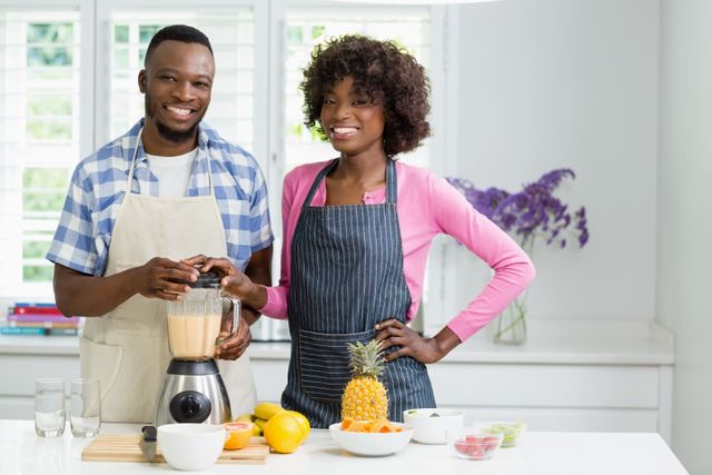 Couple in modern kitchen preparing a strawberry smoothie together. They are smiling and enjoying the process, surrounded by fresh fruits like pineapple and citrus. Ideal for use in articles or advertisements about healthy living, cooking at home, or couple activities.