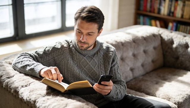Reading man on tufted sofa holding smartphone and turning page in cozy natural light