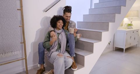 African american couple relaxing on modern staircase holding mugs smiling cozy home