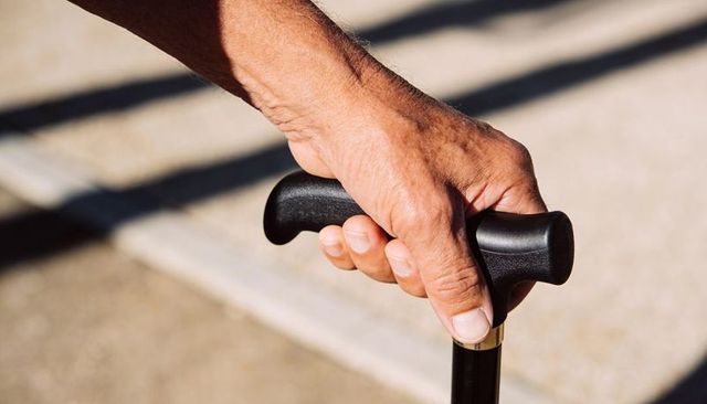 Gripping senior hand on ergonomic cane handle on sunlit sidewalk with rail shadows