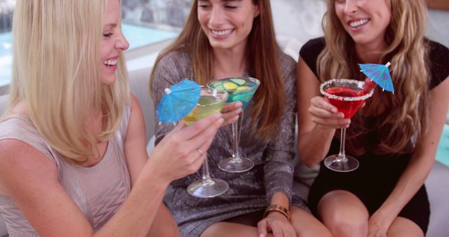 Three young women sit by the poolside lounge, holding colorful cocktails with umbrellas. They are smiling and engaging in conversation, creating a relaxed and joyful summer moment. This image is perfect for illustrating themes of friendship, social gatherings, or summer leisure. Use it for advertisements for bars, clubs, summer events, or lifestyle blogs focusing on bonding and fun experiences.