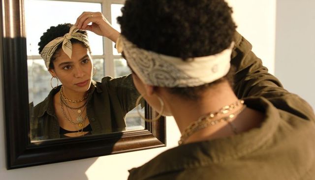Woman adjusting bandana and layering gold necklaces while checking mirror at sunlit window