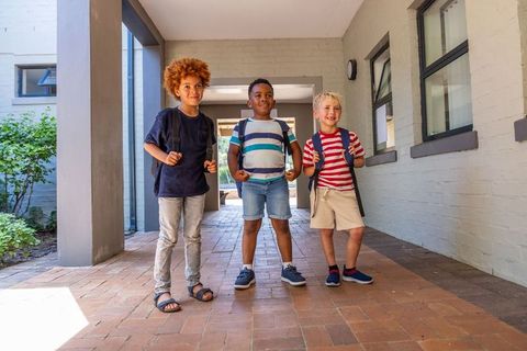 Multiracial elementary boys standing together with backpacks in sunny school corridor