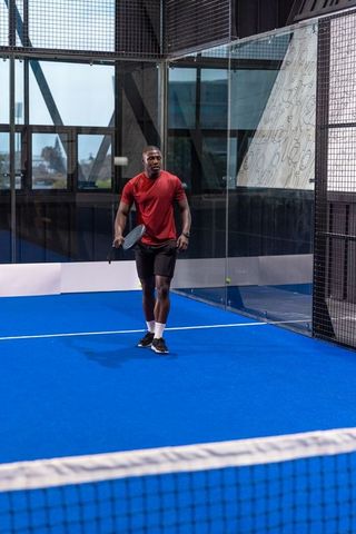 African american man playing padel on bright blue court wearing red shirt and black shorts