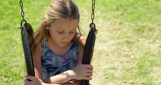 Caucasian girl sits on a swing at an outdoor park. She appears contemplative while enjoying a sunny day on the playground.