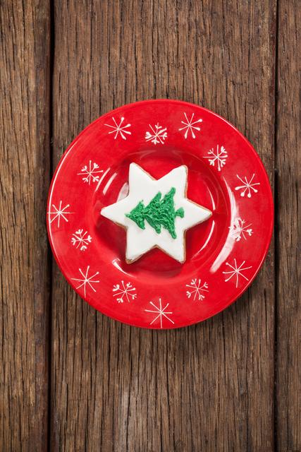 Christmas star-shaped cookie with white icing and green Christmas tree design on red plate with snowflake patterns. Wooden table background. Ideal for holiday greeting cards, festive food blogs, Christmas recipe illustrations, and seasonal marketing materials.