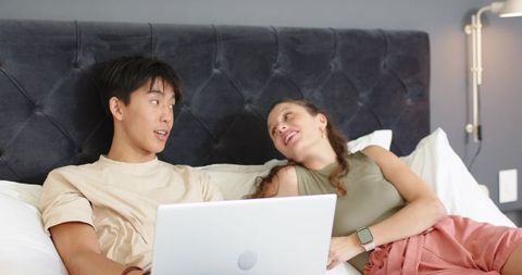 Young couple relaxing on bed sharing laptop, smiling in cozy modern bedroom lifestyle