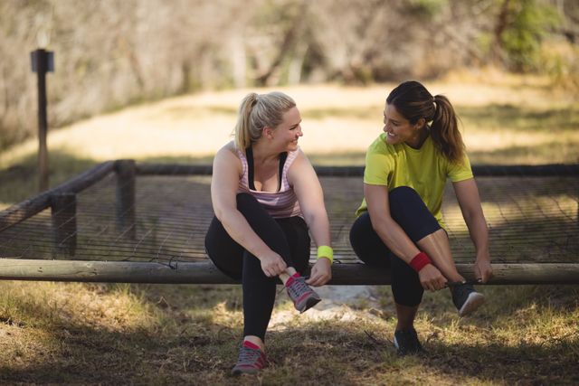 Two women are sitting on a wooden structure, putting on their shoes after completing an obstacle course workout in a boot camp. They are smiling and engaging in conversation, showcasing friendship and bonding. This image is ideal for promoting fitness, outdoor activities, teamwork, and healthy living.