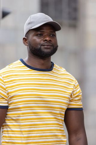 African man standing in city wearing yellow striped t-shirt and gray cap, casual urban portrait