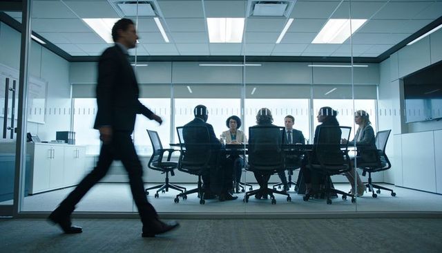 Executive team meeting behind glass conference room, business leader walking past hallway