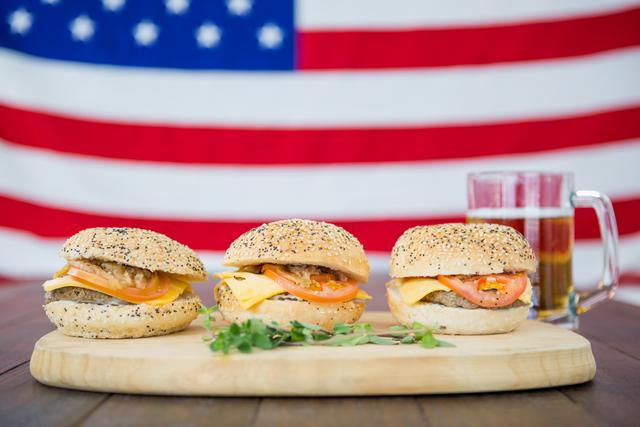 Three delicious cheeseburgers with a mug of beer on a wooden table, set against the backdrop of the USA flag. Perfect for themes related to American cuisine, patriotic celebrations, summer barbecues, Independence Day, and outdoor gatherings. Ideal for use in food blogs, restaurant promotions, holiday advertisements, and social media posts celebrating American culture.