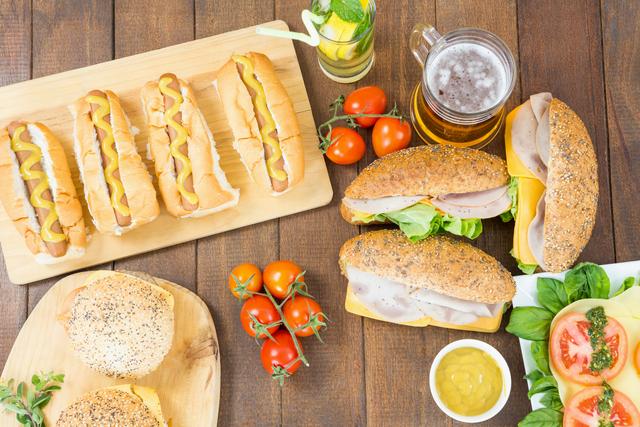 Assorted sandwiches, hot dogs, and burgers arranged on a wooden table with a glass of mojito and beer. Fresh vegetables like tomatoes and lettuce are also present, along with mustard and cheese. Ideal for use in food blogs, picnic or party invitations, and restaurant menus.