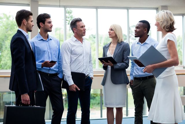 Group of business executives standing and interacting in a modern conference center lobby. They are dressed in formal attire, holding tablets and documents, engaging in a professional discussion. Ideal for use in business, corporate, teamwork, and networking contexts.
