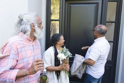 Smiling guests arriving with flowers and wine, greeting host at modern front door