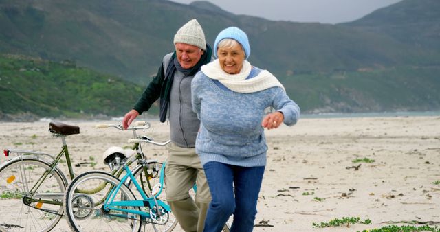 A senior Caucasian couple enjoys a leisurely walk with a bicycle on a beach, both wearing warm hats and smiling, with copy space. Their joyful expressions and casual attire suggest a moment of relaxation and happiness during a seaside retreat.