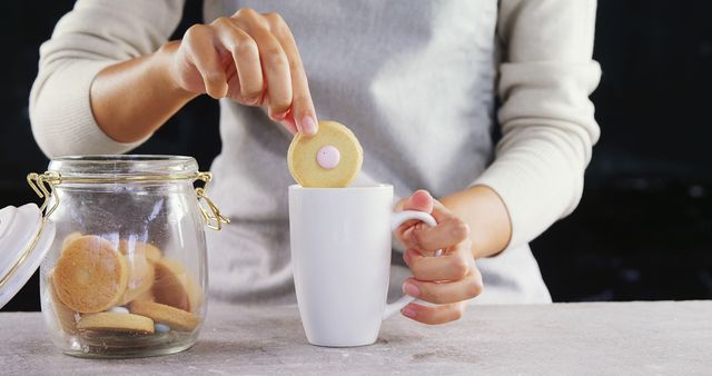 Person dipping biscuit into mug of drink with jar of cookies on table - Download Free Stock Images Pikwizard.com