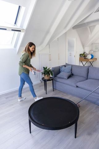 Woman dusting loft living room near skylight holding dustpan and brush in jeans