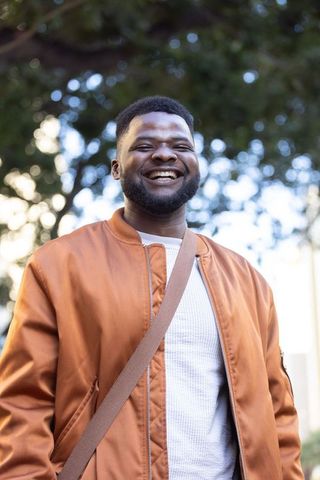 Smiling man wearing rust bomber jacket standing outdoors with crossbody bag, cheerful