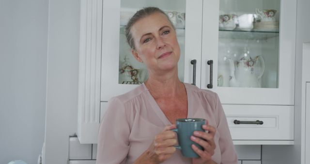 Middle-aged woman holding coffee mug in kitchen, appearing relaxed as she enjoys a moment of quiet contemplation. Suitable for themes of home comfort, morning routines, self-care, and domestic life. Great for use in advertisements and articles focused on lifestyle, health, and well-being.