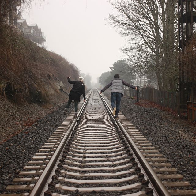 Two friends are balancing on railroad tracks on a foggy day, possibly during autumn. Bare trees and misty atmosphere create a serene and adventurous backdrop. Great for themes of friendship, adventure, autumn activities, and outdoor exploration.