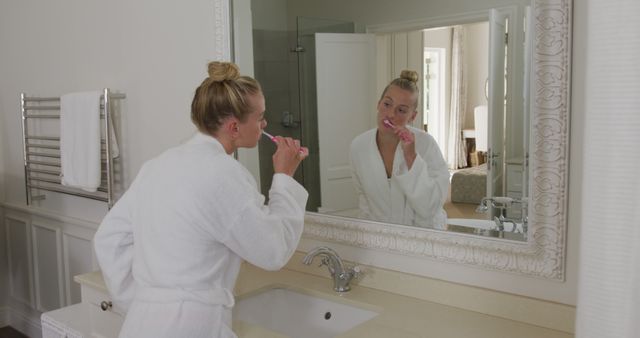 Caucasian woman in bathrobe brushing her teeth while looking in the mirror in the bathroom. hygiene and self care concept.