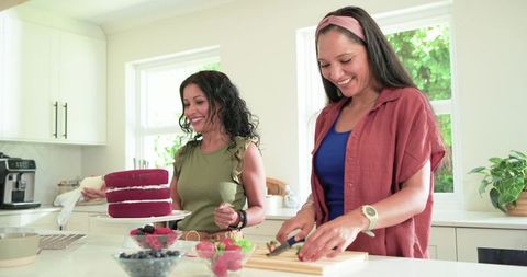 Friends baking red velvet cake together in bright home kitchen, decorating with fresh berries