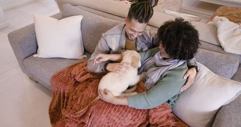Happy african american couple cuddling golden retriever puppy on sofa under cozy blanket