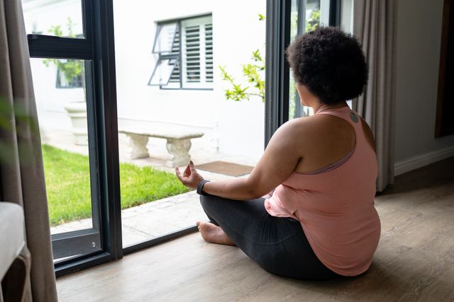 African American woman sitting cross-legged at home entrance, meditating and practicing yoga. Ideal for use in wellness, fitness, and mindfulness content, promoting healthy living and relaxation techniques.