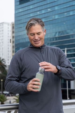 Middle-aged man opening reusable water bottle on city promenade with earbuds, smartwatch