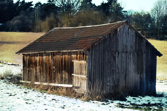 Snow-covered wooden shed standing in rural countryside field during winter. Ideal for depicting rustic charm, nostalgia, and serene natural landscapes. Perfect for winter-themed projects, rural living blogs, and agricultural content.