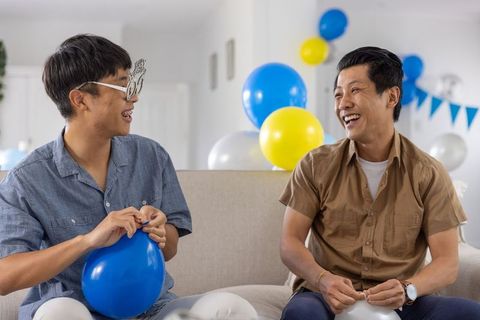 Asian men celebrating at home tying balloons on sofa smiling with party decorations
