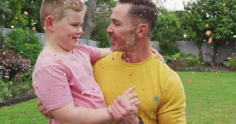 Father Joyfully Holding Son with Rainbow Confetti Celebration Outdoors