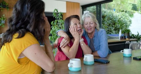 Intergenerational friends sharing emotional hug in bright kitchen with mugs, plants and smartphones