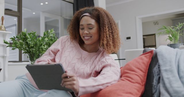 Young woman using a tablet while relaxing on a couch in living room. Ideal for articles and ads about home entertainment, online shopping, technology use, or leisure activities.