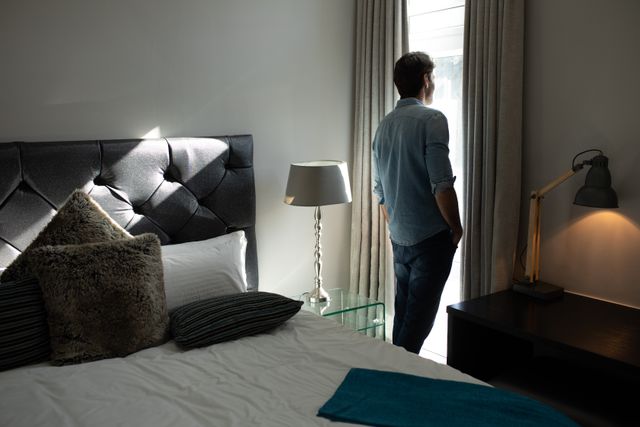 This image depicts a Caucasian man standing by the window in an elegant hotel room, looking out on a sunny day. The room features a stylish bed with pillows, a bedside lamp, and a modern desk lamp. This image can be used for travel websites, hotel advertisements, lifestyle blogs, or articles about relaxation and leisure.
