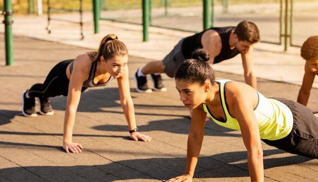 Group doing pushups outdoors on rubber tiles with bars, woman in yellow tank leading