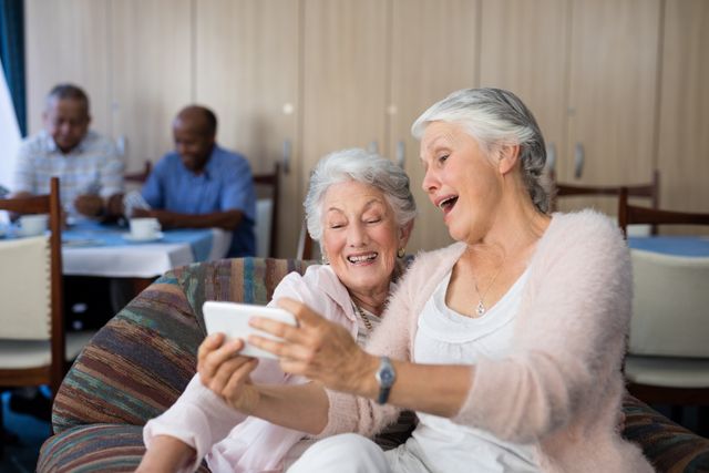 Two elderly women are sitting on a sofa in a nursing home, taking a selfie with a smartphone. They are smiling and appear to be enjoying their time together. In the background, other seniors are seen socializing at a table. This image can be used to depict themes of friendship, elderly care, technology use among seniors, and life in a retirement home.