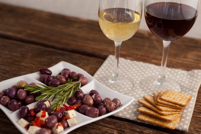 High angle view of a rustic table featuring a plate of assorted olives, cheese, and rosemary, accompanied by glasses of red and white wine and a stack of crackers. Ideal for use in food and drink blogs, gourmet dining promotions, Mediterranean cuisine advertisements, or social media posts about healthy eating and entertaining.