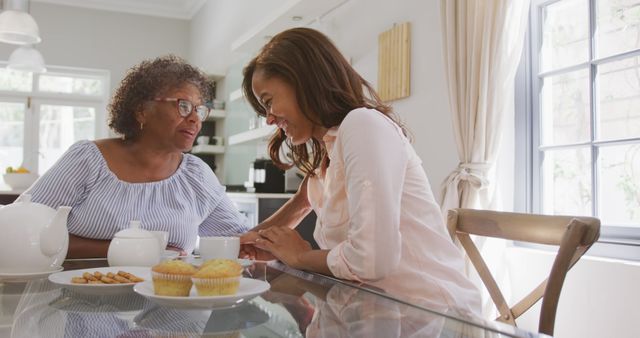 Grandmother and granddaughter sharing a happy moment at a kitchen table, drinking tea and talking. Perfect for use in family-oriented articles, social media posts celebrating family bonds, and advertising family services or home products.