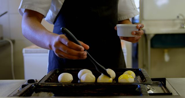 Baker in professional kitchen applying egg wash on prepared dough balls using baking brush. Ideal for content related to culinary arts, pastry baking tutorials, kitchen preparations, bakery operations, or chef training programs.