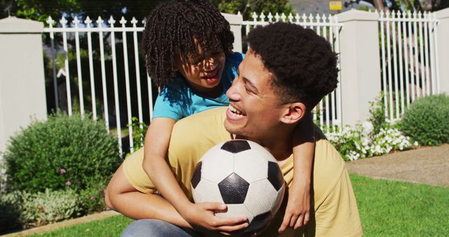 Portrait of happy biracial man and his son playing football in garden. domestic life, spending time at home.