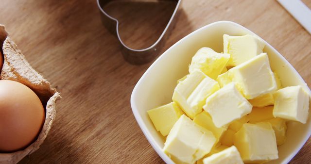 Cubed butter sits in a white bowl next to a brown egg and a cookie cutter on a wooden surface, with copy space. Ingredients are prepared for baking, suggesting the beginning of a recipe or cooking process.