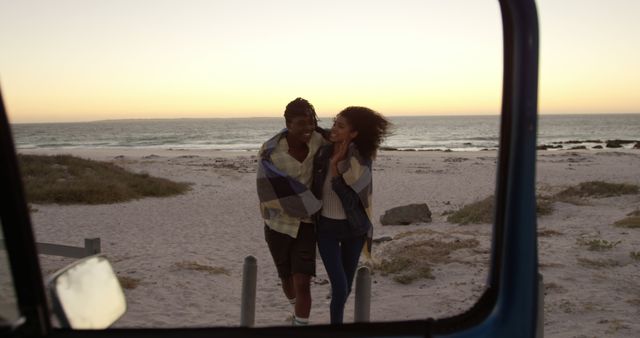 Happy african american couple walking and embracing under blanket on beach with copy space at sunset. Summer, vacations, free time, relaxing, togettherness and lifestyle, unaltered.