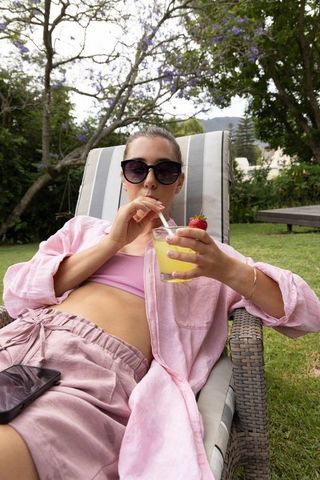Relaxing woman in pink loungewear sipping strawberry drink on chaise lounge in garden