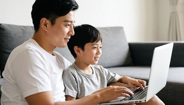 Asian father guiding son on laptop at home sofa for bonding and digital learning together