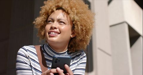 Young woman holding smartphone smiling by modern concrete facade in sunlight